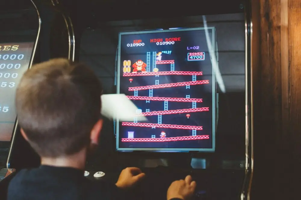 Ein Spielautomat mit Röhrenbildschirm in einer Halle, auf dem ein Spiel mit einfachen farbigen Figuren läuft; davor eine Person am Bedienfeld, unscharf.