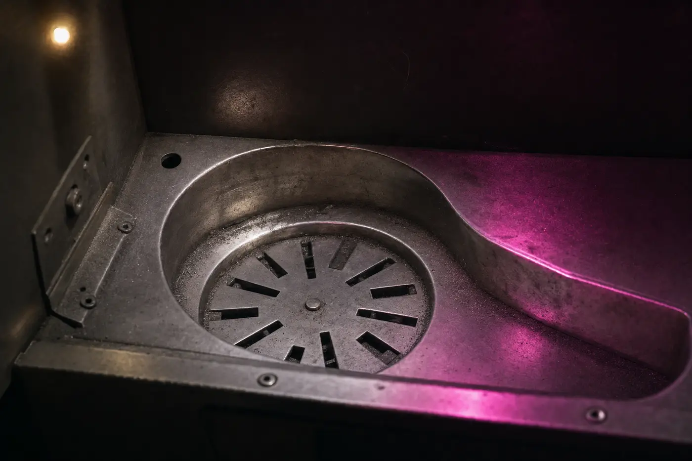 Der leere Auszahlungsbehälter eines Münzautomaten von oben: eine abgenutzte Stahlschale mit geschlitzter Drehscheibe am Boden und einer gebogenen Auswurfrinne, blank gescheuertes Metall, in den Fugen etwas grauer Staub.