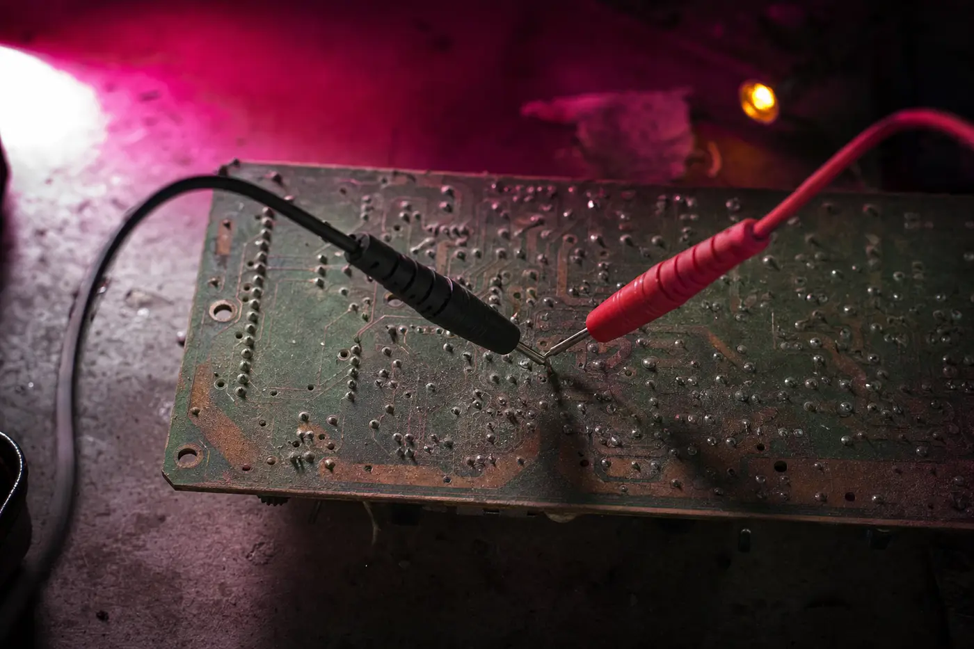 Zwei Messspitzen eines Multimeters berühren die Lötseite einer alten Platine, Makro; die rote und die schwarze Leitung queren das Bild, der Aufdruck der Platine ist bis zur Unlesbarkeit abgerieben.