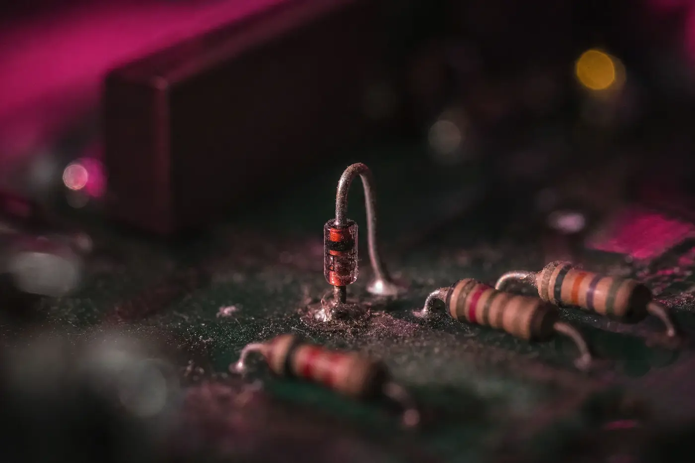 Extreme Nahaufnahme einer einzelnen kleinen Diode mit Glaskörper, aufrecht auf eine dunkelgrüne Platine gelötet, daneben einige Widerstände; ein Anschlussdraht ist leicht verbogen, im Lötlack liegt Staub.