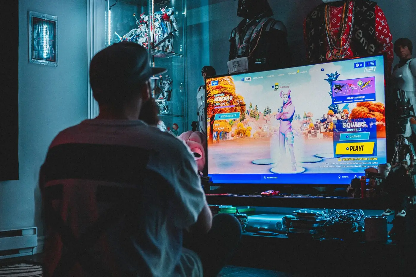 Eine Person sitzt mit dem Rücken zur Kamera vor einem Röhrenfernseher, auf dem ein farbiges Spiel läuft; der Raum ist abgedunkelt.