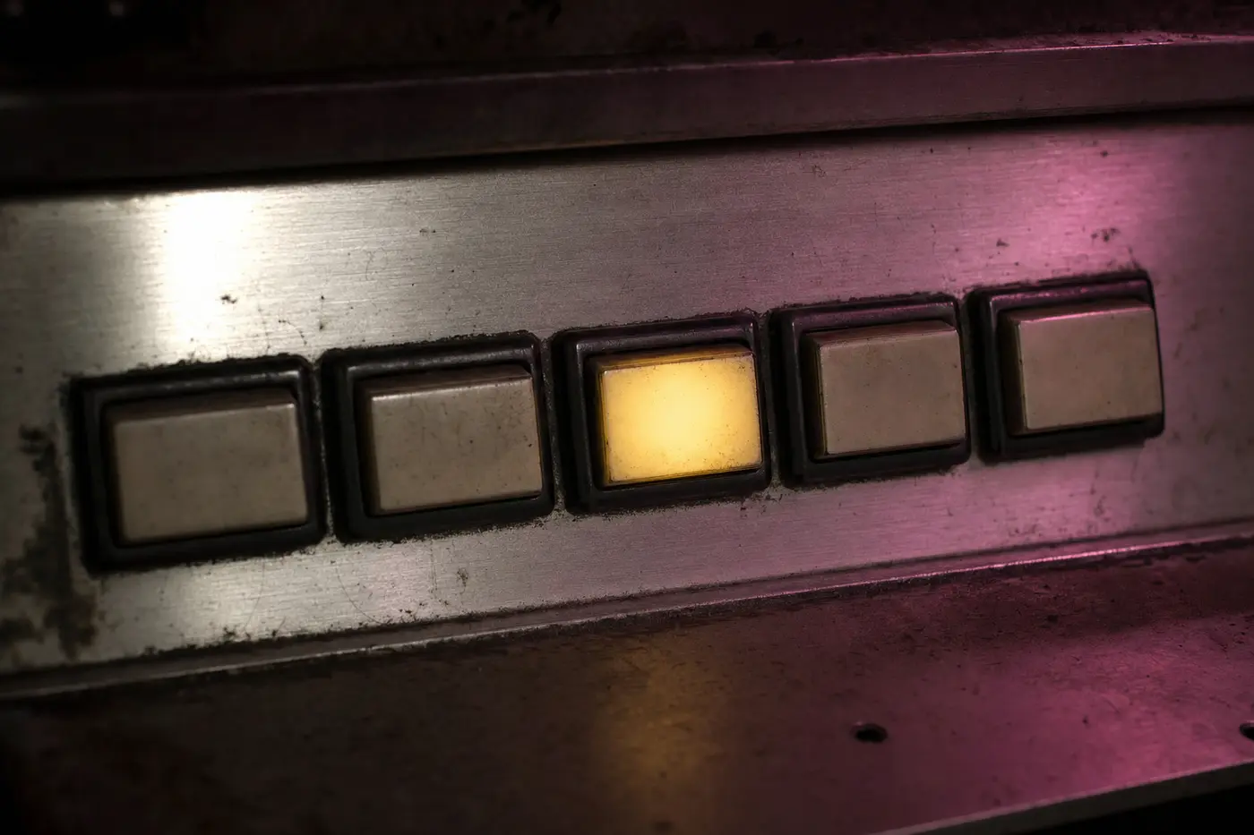 Eine Reihe von fünf abgenutzten, völlig unbeschrifteten Drucktasten auf einer gebürsteten Metallblende; eine davon leuchtet von innen warm gelb, die übrigen sind dunkel.